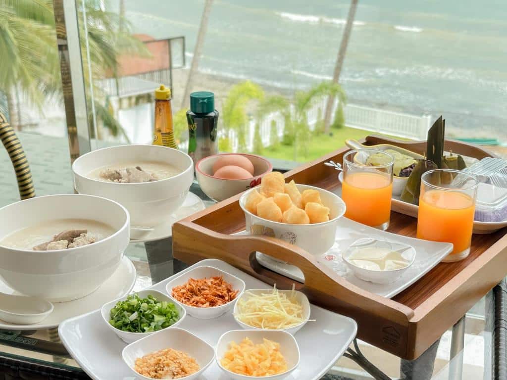 อาหารเช้าวางอยู่บนโต๊ะกระจกที่รีสอร์ทขนอม ประกอบด้วยโจ๊กข้าว ไข่ ขนมปัง เนย น้ำส้ม และเครื่องปรุงรสต่างๆ ทั้งหมดนี้เพลิดเพลินไปกับวิวทะเลที่มีต้นปาล์มและน้ำทะเลระยิบระยับ.