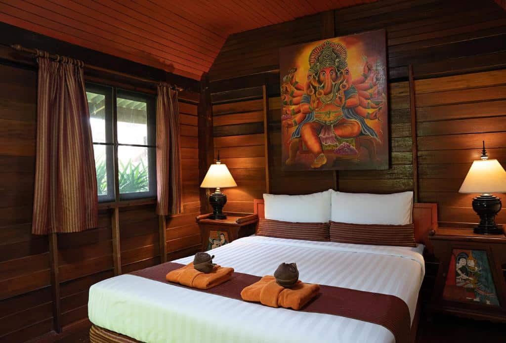 ห้องนอนไม้ที่อบอุ่นที่ ขนอมที่พัก พร้อมเตียงคู่ โคมไฟสองดวงบนโต๊ะข้างเตียง ผ้าเช็ดตัวที่พับเรียบร้อย และภาพวาดขนาดใหญ่ของพระพิฆเนศเหนือหัวเตียง.