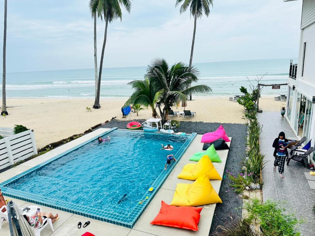 สระว่ายน้ำรูปสี่เหลี่ยมผืนผ้าที่มีผู้คนกำลังว่ายน้ำ ถุงฟองน้ำสีสันสดใสอยู่ข้างๆ ต้นปาล์ม และชายหาดทรายพร้อมคลื่นทะเลในพื้นหลังที่รีสอร์ทขนอม.