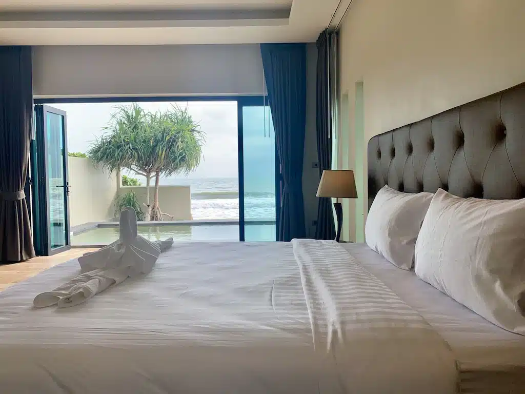 เตียงที่จัดไว้อย่างเรียบร้อยหันหน้าไปทางประตูกระจกบานใหญ่ เปิดออกสู่ลานระเบียงที่มีสระว่ายน้ำ ต้นปาล์ม และวิวทะเล—เหมาะสำหรับการเข้าพักของคุณที่ ที่พักขนอม ผ้าขนหนูพับเป็นรูปหงส์วางอยู่บนเตียง.