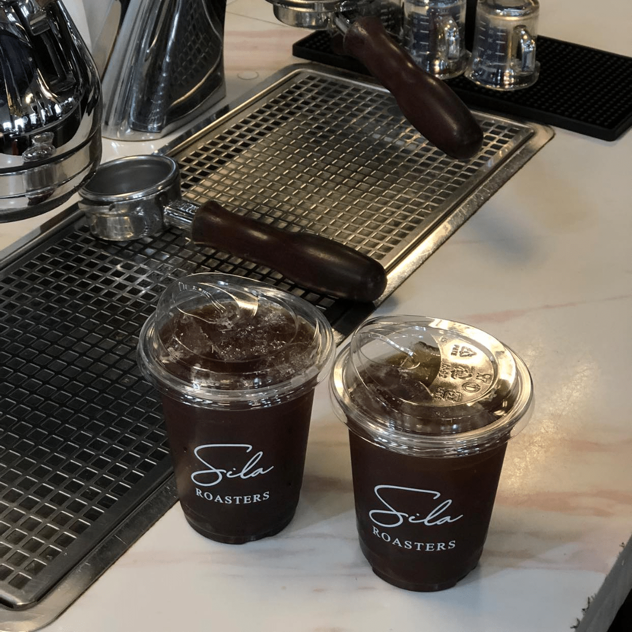 เครื่องดื่มกาแฟเย็นสองแก้วในแก้วพลาสติกใสที่มีฉลาก "Sila Roasters" ตั้งอยู่บนเคาน์เตอร์ข้างๆ เครื่องชงเอสเพรสโซ่—เครื่องดื่มที่สดชื่นสมบูรณ์แบบก่อนการผจญภัยครั้งต่อไปของคุณไปยังสถานที่ท่องเที่ยวธรรมชาติใกล้กรุงเทพฯ หรือ น้ำตกใกล้กรุงเทพฯ.