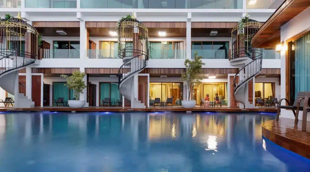 โรงแรมสมัยใหม่ ณ โรงแรมขนอม พร้อมห้องพักที่หันหน้าเข้าสระว่ายน้ำ โดดเด่นด้วยบันไดวน ระเบียง หน้าต่างบานใหญ่ และต้นไม้กระถางบนดาดฟ้า.