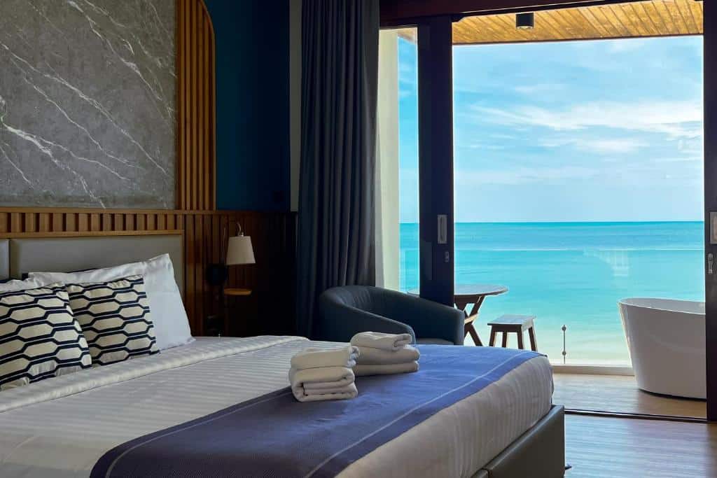 ห้องพักโรงแรมที่รีสอร์ทขนอม พร้อมเตียงที่จัดไว้อย่างเรียบร้อย ผ้าเช็ดตัววางซ้อนกัน เก้าอี้นวม อ่างอาบน้ำ และหน้าต่างบานใหญ่ที่มองเห็นทะเลสีฟ้าและท้องฟ้าที่สงบเงียบ.