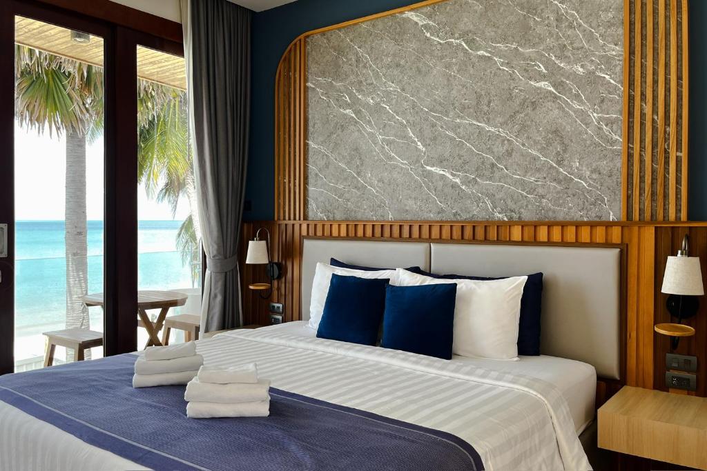 ห้องพักโรงแรมขนอมที่ทันสมัยพร้อมเตียงที่จัดไว้อย่างเรียบร้อย ผ้าเช็ดตัวพับไว้อย่างเรียบร้อย ตกแต่งด้วยสีน้ำเงิน หัวเตียงหินอ่อน และหน้าต่างบานใหญ่ที่มองเห็นชายหาดและต้นปาล์ม.