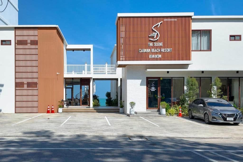 รีสอร์ทริมชายหาดสมัยใหม่พร้อมด้วยไม้และสีขาวภายนอก ป้ายเขียนว่า "The Scene Cabana Beach Resort" รถยนต์จอดอยู่ พืชพรรณเขียวชอุ่ม และที่จอดรถว่างใต้ท้องฟ้าแจ่มใส—เหมาะสำหรับผู้ที่มองหา โรงแรมขนอม หรือ ที่พักขนอม.