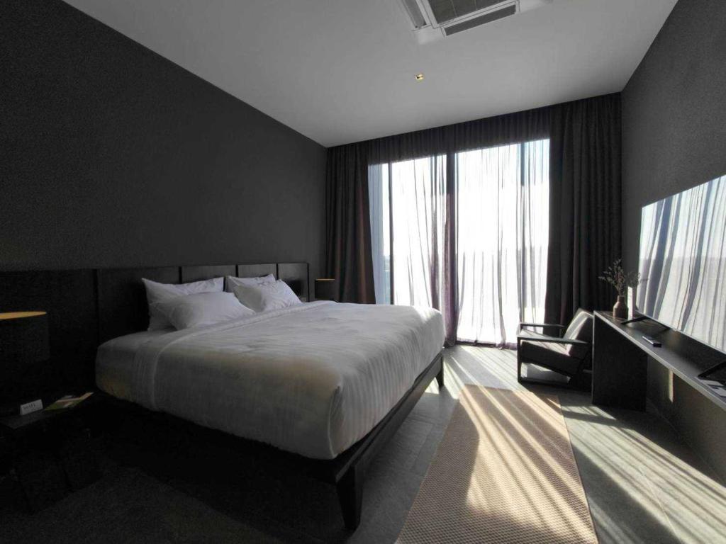 ห้องพักโรงแรมสมัยใหม่ที่ โรงแรมเพชรบูรณ์ ประกอบด้วยเตียงขนาดใหญ่ ผนังสีเข้ม โต๊ะทำงาน และเก้าอี้ พร้อมแสงแดดส่องผ่านผ้าม่านบางที่ปิดหน้าต่างจากพื้นจรดเพดาน.