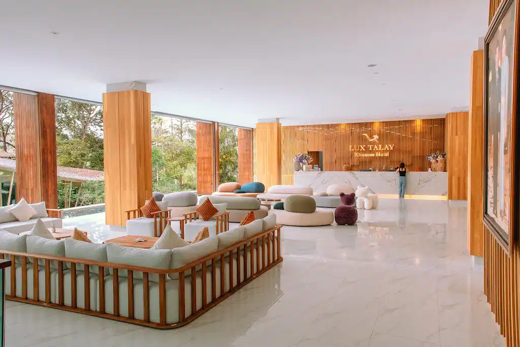 ล็อบบี้โรงแรมสมัยใหม่ตกแต่งด้วยไม้ พื้นหินอ่อน โซฟาสีขาว หมอนสีสันสดใส และเคาน์เตอร์ต้อนรับที่มีพนักงานยืนอยู่ใต้ป้ายที่เขียนว่า "Lux Talay Khanna Hotel" หนึ่งในโรงแรมชั้นนำในขนอมสำหรับการเข้าพักของคุณ.