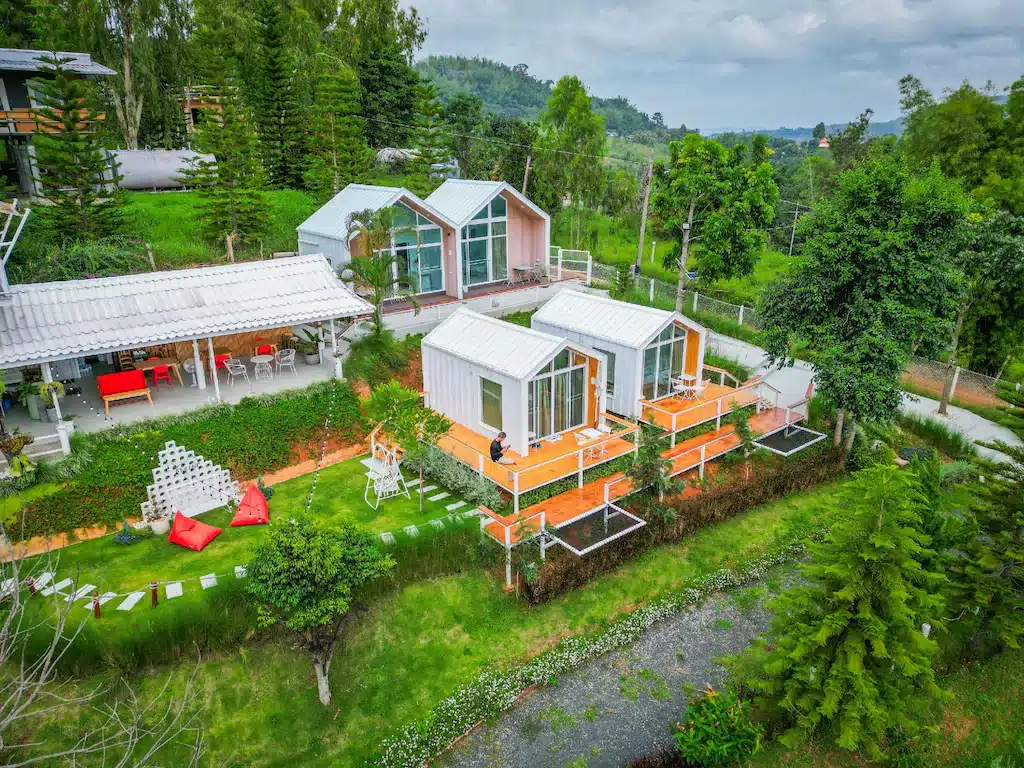 มุมมองจากอากาศของบ้านเล็กสมัยใหม่ที่มีหน้าต่างขนาดใหญ่และเฉลียงสีส้มที่รีสอร์ทเพชรบูรณ์ ล้อมรอบด้วยต้นไม้เขียวขจีและพื้นที่นั่งเล่นกลางแจ้งในสวนที่ตกแต่งอย่างสวยงาม.