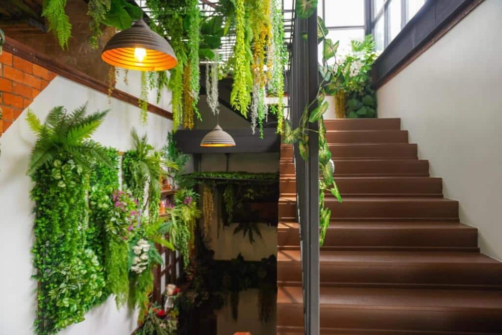 บันไดสีน้ำตาลอยู่ถัดจากผนังที่ตกแต่งด้วยพืชสีเขียวหลากหลายชนิดและเฟิร์น ภายใต้โคมไฟแขวนสองดวงในพื้นที่ภายในที่สว่างไสว ที่พักขนอมที่แสนอบอุ่น.