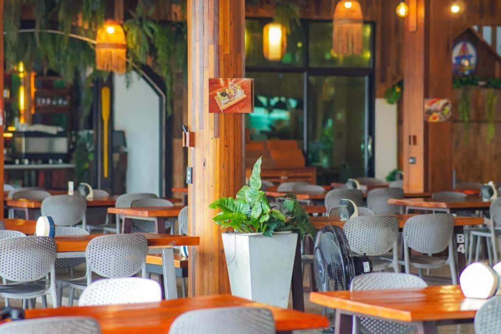 ร้านอาหารที่มีโต๊ะไม้ เก้าอี้สีเทา โคมไฟแขวน ต้นไม้ในกระถาง และพื้นที่บาร์อยู่ด้านหลัง—เหมาะสำหรับแขกที่ต้องการรับประทานอาหารอย่างมีสไตล์ที่ โรงแรมขนอม.
