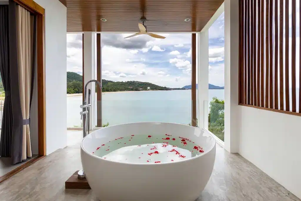 อ่างอาบน้ำสีขาวทันสมัยที่เต็มไปด้วยน้ำและกลีบดอกไม้ตั้งอยู่ข้างหน้าต่างบานใหญ่ที่มองเห็นทะเลที่สงบและภูเขาไกลๆ—ช่วงเวลาที่งดงามที่รีสอร์ทขนอมภายใต้ท้องฟ้าที่มีเมฆบางส่วน.