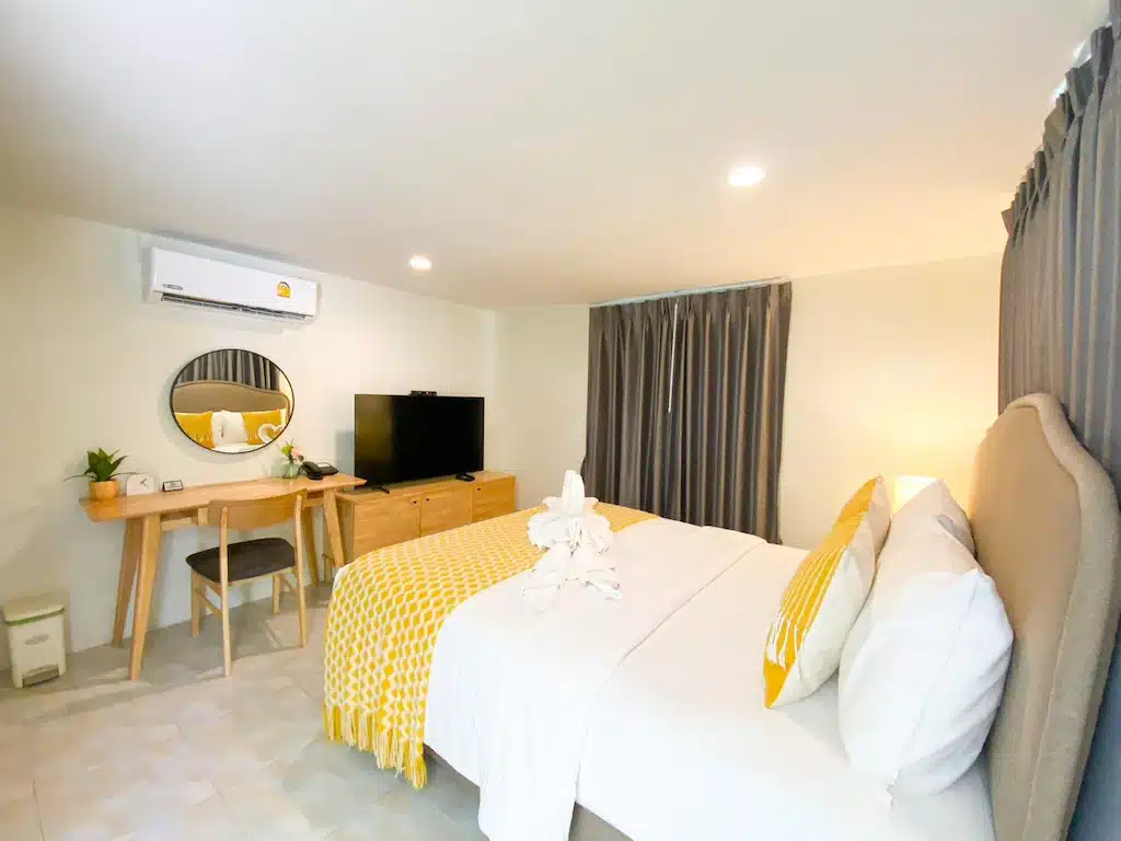 ห้องพักในโรงแรมที่ โรงแรมขนอม พร้อมเตียงคู่ ตกแต่งด้วยสีเหลือง โต๊ะทำงานพร้อมกระจกกลม ทีวีบนตู้ไม้ เครื่องปรับอากาศ และม่านปิดสนิท—เหมาะสำหรับการเข้าพักอย่างมีสไตล์ใน ขนอมที่พัก.
