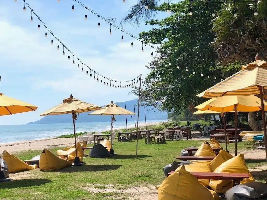 พื้นที่นั่งเล่นริมชายหาดกลางแจ้งพร้อมร่มสีเหลือง, ถุงเมล็ด, และไฟประดับที่ ที่พักขนอม, มองเห็นมหาสมุทรและภูเขาไกล ๆ ภายใต้ท้องฟ้าที่มีเมฆบางส่วน.