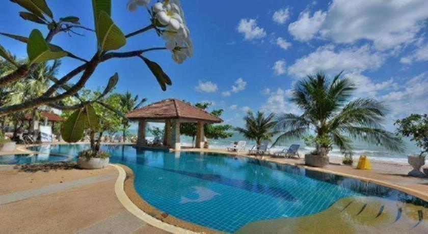 สระว่ายน้ำกลางแจ้งพร้อมศาลา ล้อมรอบด้วยต้นปาล์มและพืชเขตร้อน ณ รีสอร์ทขนอม มองเห็นชายหาดทรายและมหาสมุทรภายใต้ท้องฟ้าสีครามที่มีเมฆลอยประปราย.