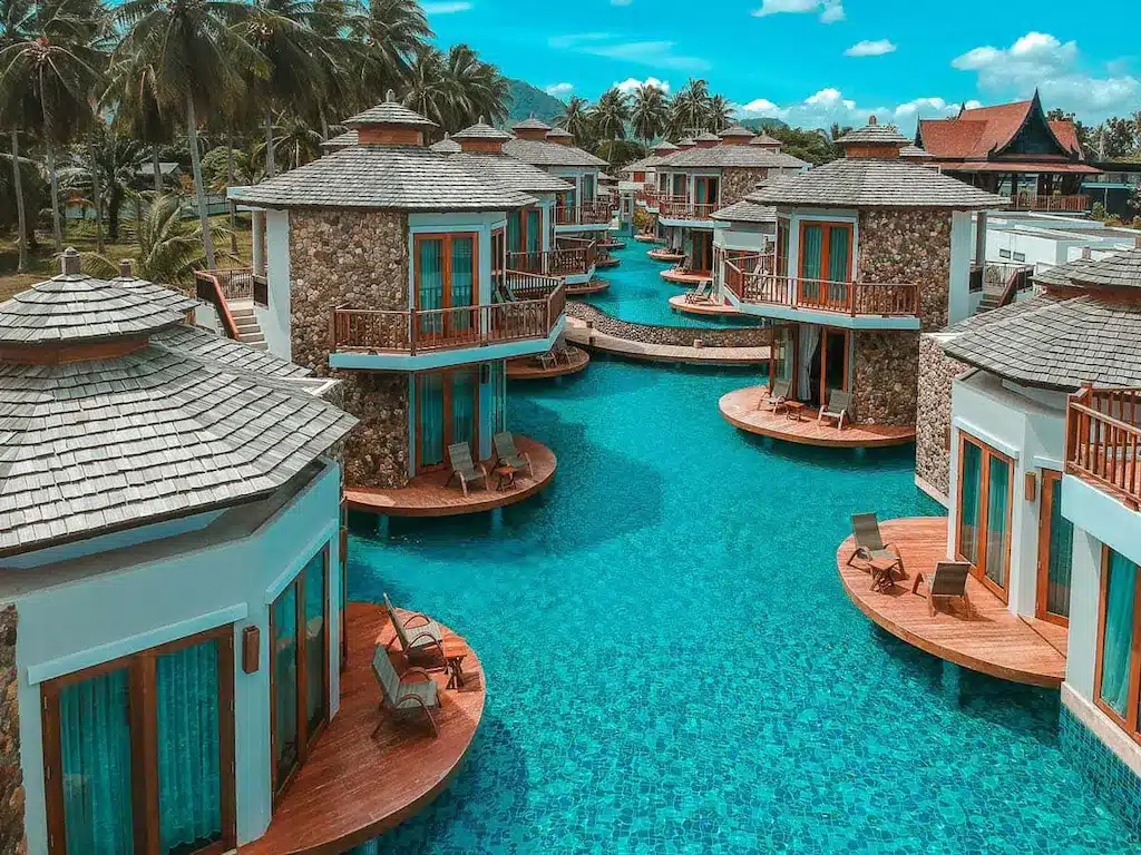 บังกะโลกลางน้ำพร้อมระเบียงส่วนตัวและเก้าอี้เลานจ์ตั้งอยู่เหนือสระว่ายน้ำสีฟ้าใสที่ที่พักในขนอมอันเงียบสงบแห่งนี้ ล้อมรอบด้วยต้นปาล์มและภูเขาภายใต้ท้องฟ้าสดใส.