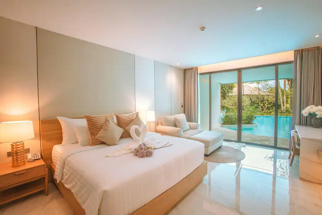 ห้องพักโรงแรมสมัยใหม่ในขนอม พร้อมเตียงคู่ โต๊ะข้างเตียง โซฟา และประตูบานกระจกขนาดใหญ่ที่เปิดออกสู่ทัศนียภาพริมสระว่ายน้ำ ผ้าเช็ดตัวบนเตียงพับเป็นรูปหัวใจ—เหมาะสำหรับการเข้าพักของคุณที่ที่พักในขนอม.