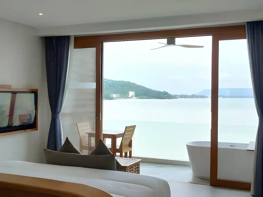 ห้องพักโรงแรมสมัยใหม่พร้อมประตูกระจกบานใหญ่เปิดออกสู่ระเบียงที่มองเห็นทะเลสงบและเนินเขาไกลๆ อ่างอาบน้ำแบบลอยตัวใกล้หน้าต่างเพิ่มความหรูหรา—เหมาะสำหรับผู้ที่มองหา รีสอร์ทขนอม หรือ ที่พักขนอม ริมน้ำที่เงียบสงบ.