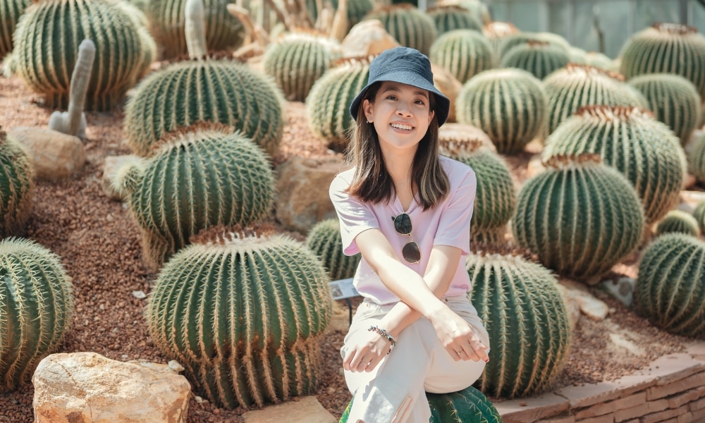 ผู้หญิงคนหนึ่งสวมหมวกแก๊ปและเสื้อผ้าสีอ่อนนั่งยิ้มอยู่ท่ามกลางกระบองเพชรกลมขนาดใหญ่ในสวนทะเลทรายที่อาบไปด้วยแสงแดด สะท้อนบรรยากาศสบายๆ ของ ที่เที่ยวม่อนแจ่ม.