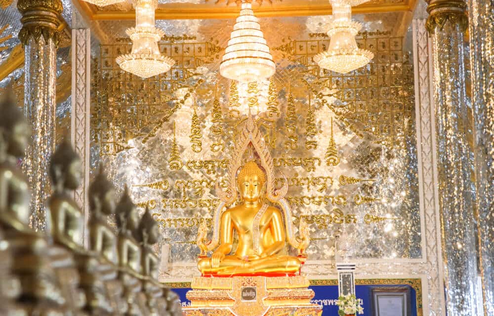 พระพุทธรูปทองคำตั้งอยู่ในห้องที่ตกแต่งอย่างวิจิตรบรรจง มีตัวอักษรทองคำและลวดลายซับซ้อนบน ที่เที่ยวใกล้กรุงเทพ ผนังและโคมระย้าที่แขวนอยู่ด้านบน—เป็นจุดแวะที่น่าประทับใจสำหรับผู้ที่ต้องการ เที่ยวใกล้กรุงเทพ.