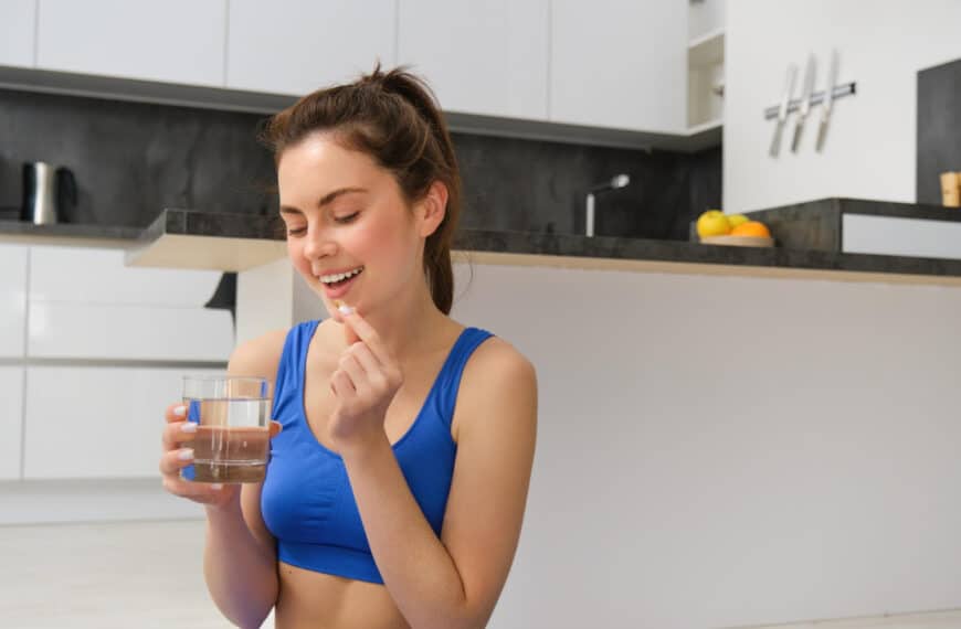 หญิงสาวในชุดออกกำลังกายถือแก้วน้ำและเตรียมจะกินยาเม็ดสีขาวในครัวสมัยใหม่.