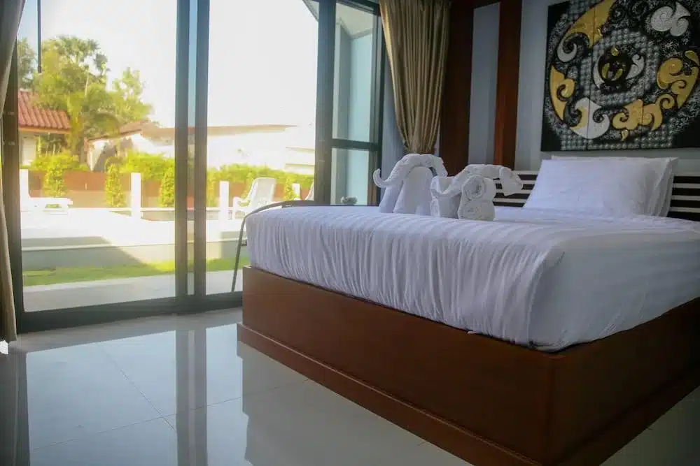 ห้องนอนสมัยใหม่พร้อมเตียงที่จัดไว้อย่างเรียบร้อย ประติมากรรมผ้าขนหนูรูปช้าง หน้าต่างบานใหญ่ และวิวของลานและสวนด้านนอก—เหมาะสำหรับผู้ที่มองหาที่พักในสไตล์ที่หรูหรา.