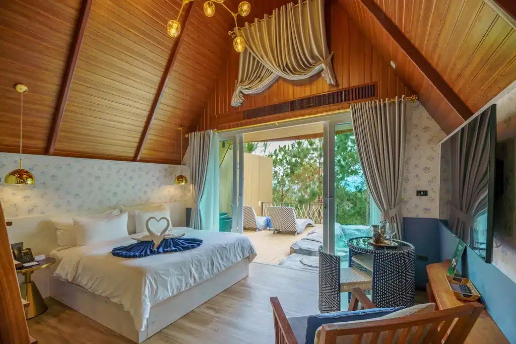 ห้องนอนที่แสนสบายที่โรงแรมเพชรบูรณ์ พร้อมเพดานไม้ลาดเอียง เตียงขนาดใหญ่ ผ้าม่านตกแต่ง ทีวีติดผนัง และประตูกระจกบานเลื่อนที่เปิดออกสู่ระเบียง เหมาะสำหรับแขกที่ต้องการที่พักในเมืองเพชรบูรณ์พร้อมวิวธรรมชาติ.