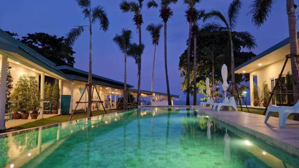 สระว่ายน้ำกลางแจ้งล้อมรอบด้วยต้นปาล์มและอาคารต่าง ๆ ส่องแสงสว่างในยามพลบค่ำ ท้องฟ้าค่อย ๆ เปลี่ยนเป็นราตรี—สถานที่พักผ่อนในบรรยากาศเงียบสงบที่รีสอร์ทขนอม เหมาะสำหรับการผ่อนคลายอย่างแท้จริง.