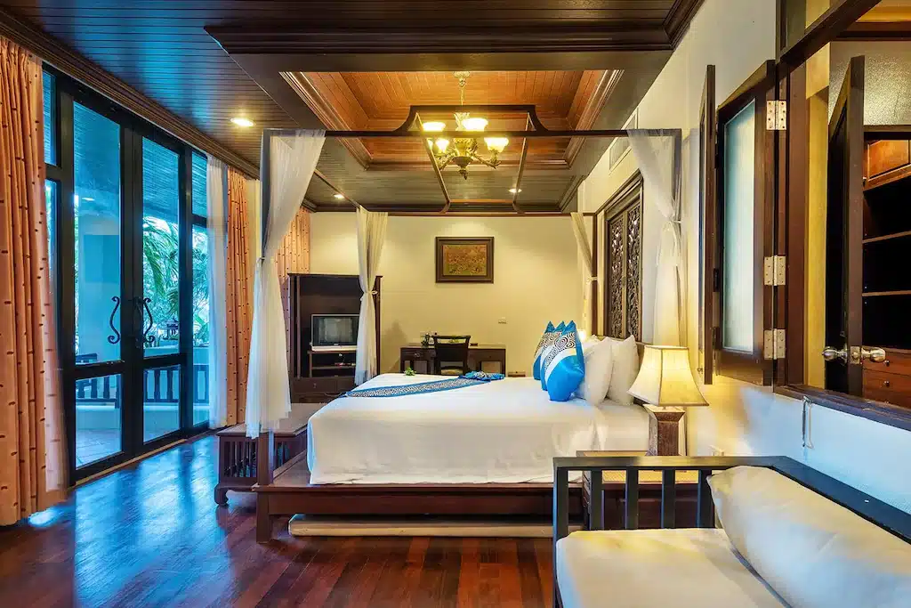 ห้องนอนกว้างขวางที่ โรงแรมขนอม พร้อมเตียงไม้สี่เสา ผ้าปูที่นอนสีขาว โซฟา พื้นไม้ หน้าต่างบานใหญ่พร้อมผ้าม่าน และแสงไฟอบอุ่น.