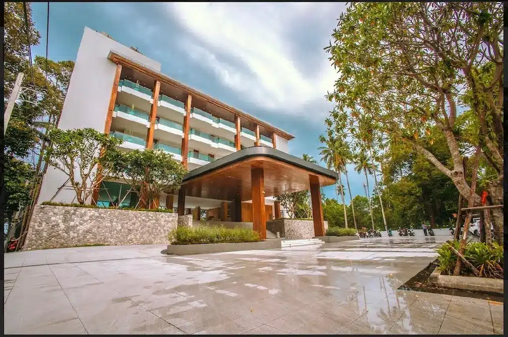 โรงแรมหลายชั้นที่ทันสมัยพร้อมระเบียงกระจกและทางเข้าที่มีหลังคาคลุม ล้อมรอบด้วยต้นไม้และทางเดินที่ปูด้วยหินในวันที่เมฆครึ้ม—เหมาะสำหรับผู้ที่มองหา รีสอร์ทขนอม ในบรรยากาศที่เงียบสงบ.
