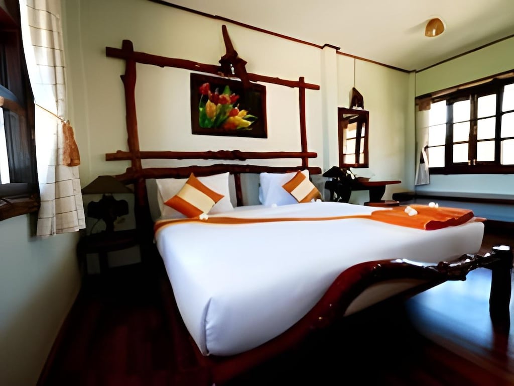 เตียงที่จัดไว้อย่างเรียบร้อยพร้อมหมอนสีส้มและขาวในห้องที่สว่างไสวด้วยเฟอร์นิเจอร์ไม้ หน้าต่างบานใหญ่ และภาพวาดดอกไม้เหนือหัวเตียง—บรรยากาศที่สมบูรณ์แบบสำหรับการพักผ่อนของคุณที่ รีสอร์ทขนอม.