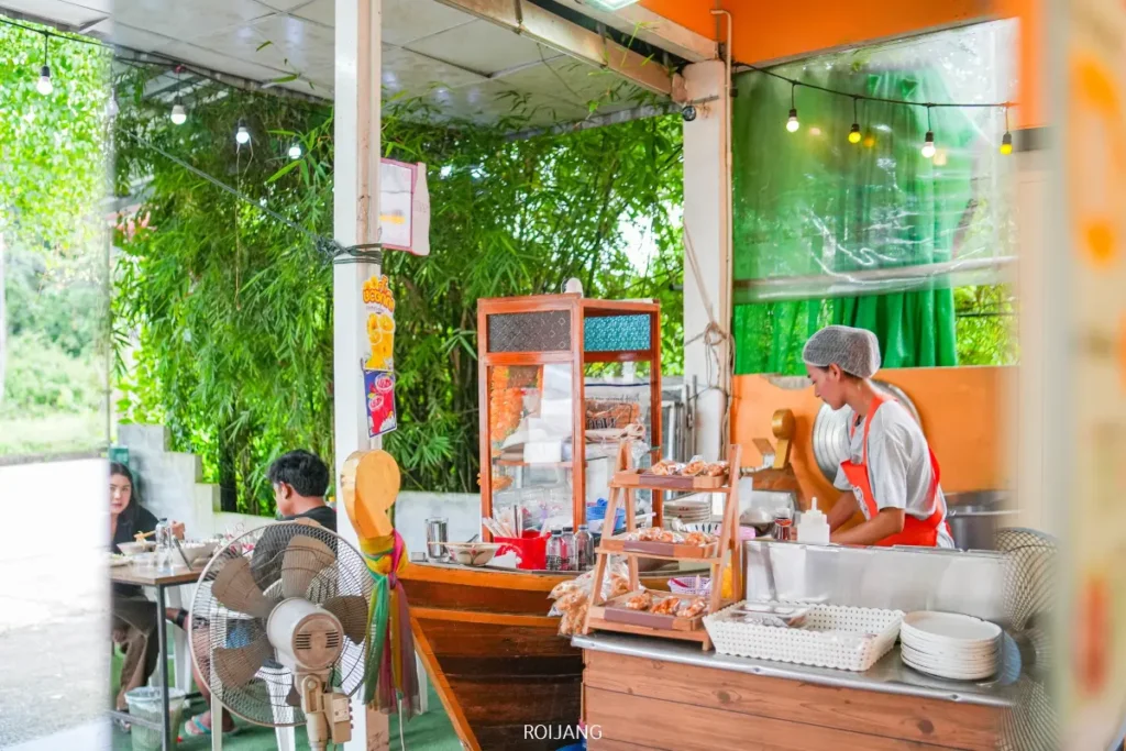 พ่อค้าในผ้ากันเปื้อนสีส้มกำลังเตรียมอาหารที่ร้านอาหารแบบเปิดโล่งชื่อ Bear Grill ซึ่งมีเคาน์เตอร์รูปเรือและของว่างหลากหลายชนิดวางโชว์อยู่ มีลูกค้าสองคนนั่งอยู่ที่โต๊ะในฉากหลัง.