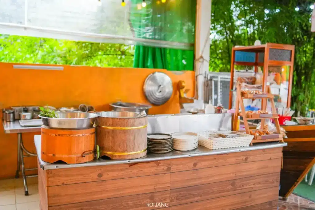 เคาน์เตอร์อาหารไม้ Bear Grill พร้อมหม้อ จาน และช้อนส้อม ตั้งอยู่ในครัวกลางแจ้งที่มีผนังสีส้มและต้นไม้สีเขียวเป็นฉากหลัง.