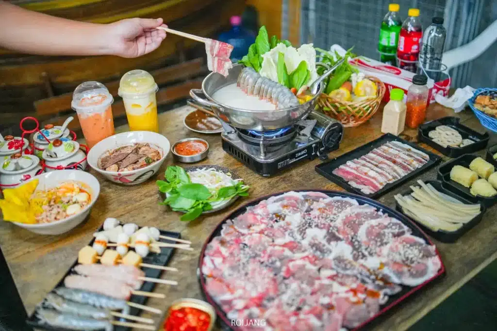 การเสิร์ฟแบบแบร์กริลล์บนโต๊ะประกอบด้วยเนื้อดิบ ผัก ซอส และเครื่องดื่มที่วางล้อมรอบหม้อไฟร้อน โดยมีคนจุ่มเนื้อหั่นบางลงในน้ำซุปที่กำลังเดือด.