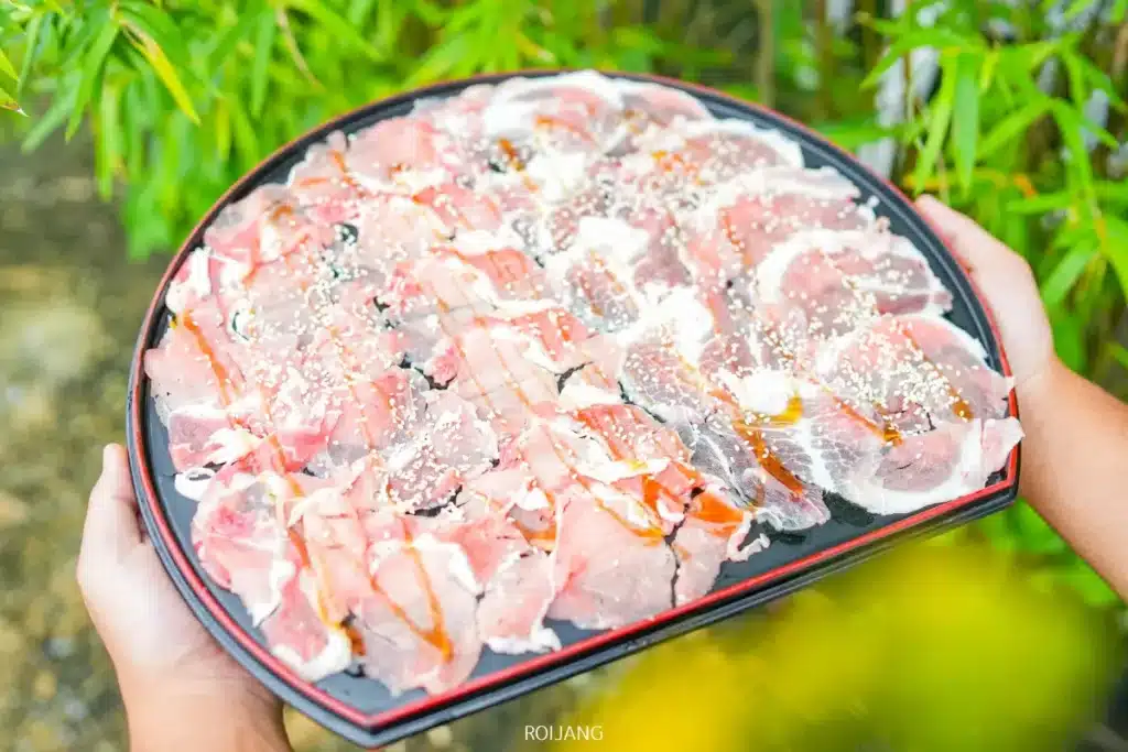 บุคคลหนึ่งถือถาด Bear Grill ขนาดใหญ่ทรงกลมที่เต็มไปด้วยเนื้อสดหั่นบางๆ โรยด้วยงาดำ โดยมีพืชสีเขียวอยู่ด้านหลัง.