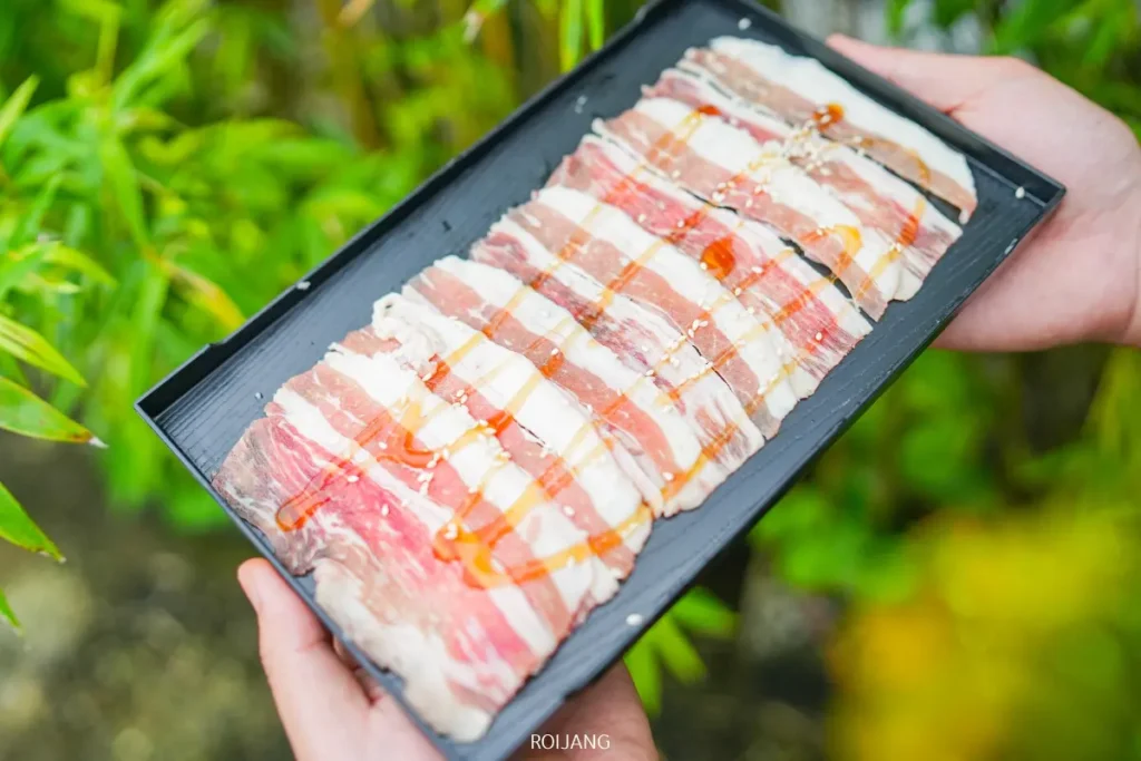 ถาดสีดำจาก Bear Grill วางชิ้นเนื้อหมูสามชั้นสดที่มีลายมันแทรก ราดด้วยซอสเล็กน้อย โดยมีพืชสีเขียวเป็นฉากหลัง.