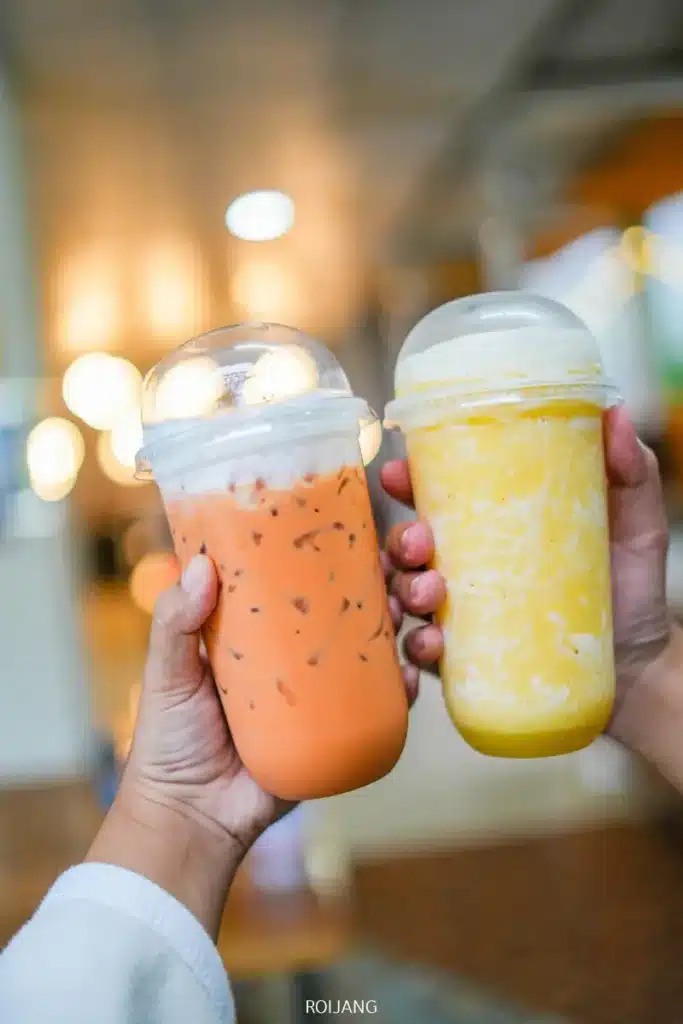 สองมือถือแก้วพลาสติก Bear Grill ที่มีฝาโดมปิดอยู่ หนึ่งแก้วเต็มไปด้วยชาไทยเย็นและอีกแก้วเต็มไปด้วยสมูทตี้สีเหลือง วางอยู่ต่อหน้าฉากหลังภายในที่เบลอ.