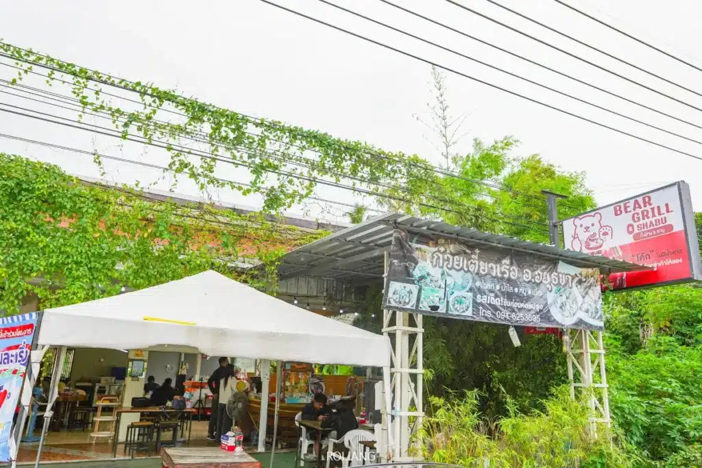 วิวภายนอกของร้านอาหาร Bear Grill พร้อมลูกค้าที่นั่งอยู่ใต้เต็นท์สีขาว ท่ามกลางต้นไม้เขียวชอุ่ม และมีป้าย Bear Grill ที่โดดเด่นอยู่เหนือศีรษะ.