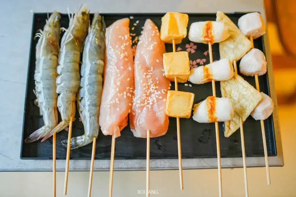 ถาดย่างแบล็กแบร์วางเสียบกุ้งสด ไก่หน้าอกคลุกงา เต้าหู้หั่นเต๋า และปลาแผ่นหลากชนิดเรียงกันเป็นแถว.