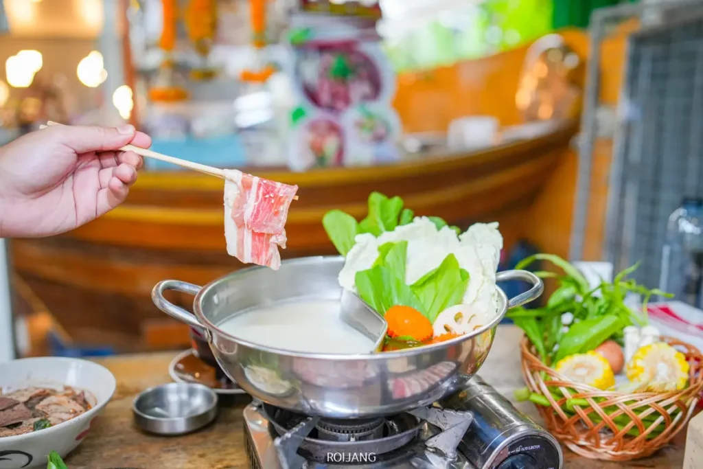 มือหนึ่งถือเนื้อหั่นบางๆ อยู่เหนือหม้อ Bear Grill ที่มีน้ำซุปเดือดปุดๆ พร้อมผักและเต้าหู้บนโต๊ะ พร้อมสำหรับการรับประทานหม้อไฟ.