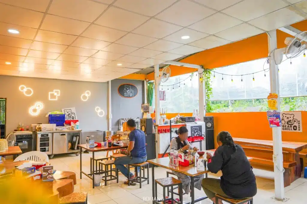 ร้านอาหารขนาดเล็กที่สว่างไสวอย่างสวยงามชื่อ Bear Grill มีผู้คนไม่กี่คนนั่งรับประทานอาหารและคุยกันอยู่ที่โต๊ะ ผนังสีส้ม, ศิลปะบนผนัง, และหน้าต่างขนาดใหญ่ทำให้พื้นที่เต็มไปด้วยแสงสว่างจากธรรมชาติ.