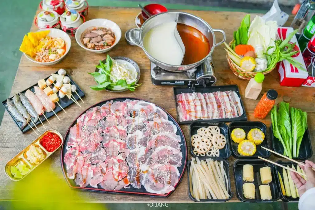 อาหารหม้อไฟร้อนบนโต๊ะไม้ประกอบด้วยเนื้อสไลซ์ ผัก ซอสจิ้ม ไม้เสียบ และหม้อ Bear Grill ที่มีน้ำซุปสองชนิดกำลังเคี่ยวบนเตาพกพา.