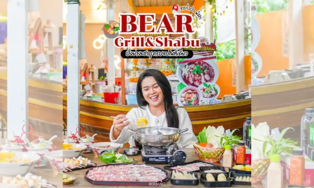 บุคคลหนึ่งนั่งอยู่ที่โต๊ะพร้อมด้วยวัตถุดิบสำหรับชาบูชาบูและปิ้งย่างหลากหลายชนิด ยิ้มและกำลังปรุงอาหารที่ร้าน Bear Grill & Shabu.