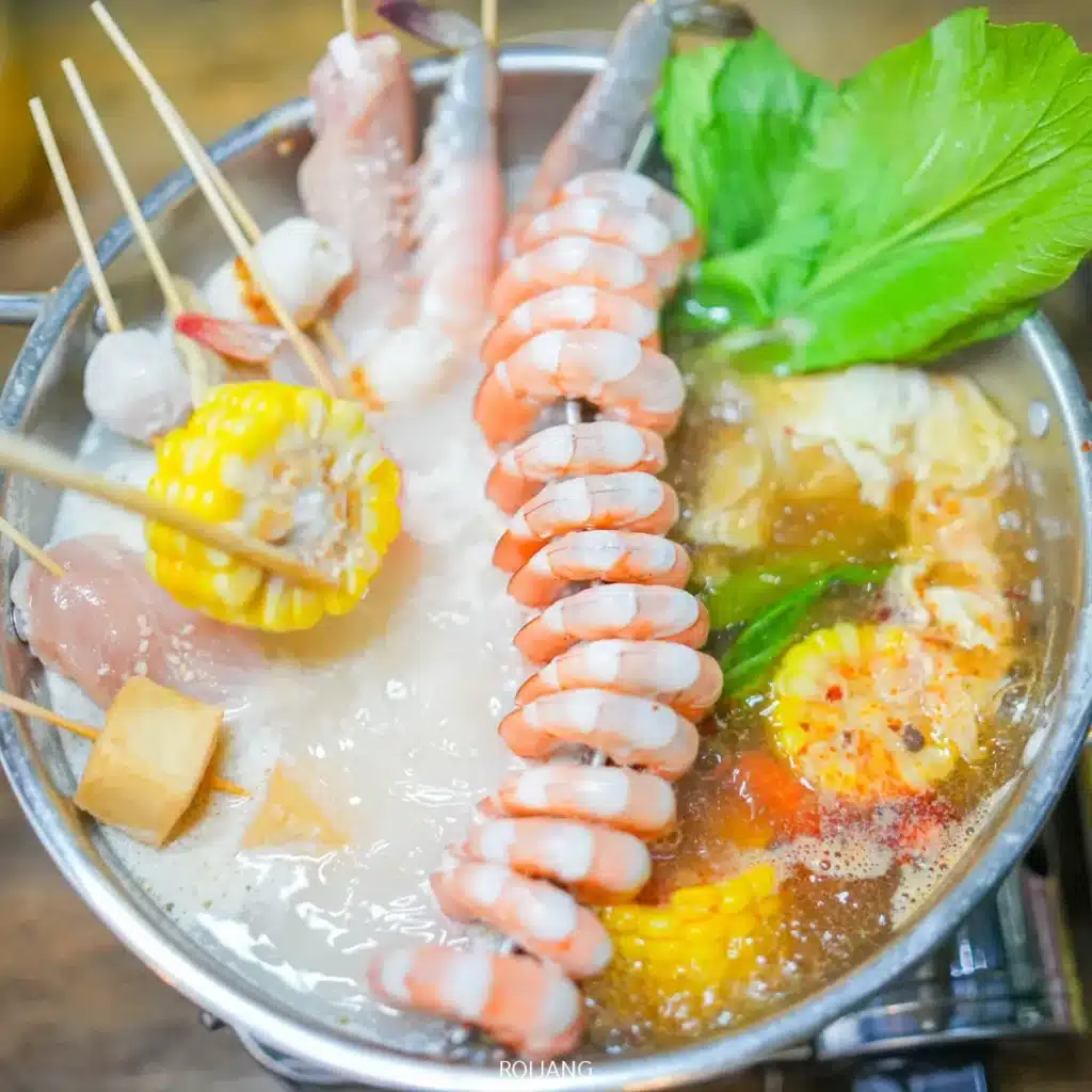 หม้อหุงต้มแบบบาร์บีคิวที่มีน้ำซุปเดือดปุด ๆ พร้อมกุ้ง ข้าวโพด ผักใบเขียว เนื้อไม้เสียบ เต้าหู้ และผักต่าง ๆ ที่กำลังปรุงอยู่ภายใน.