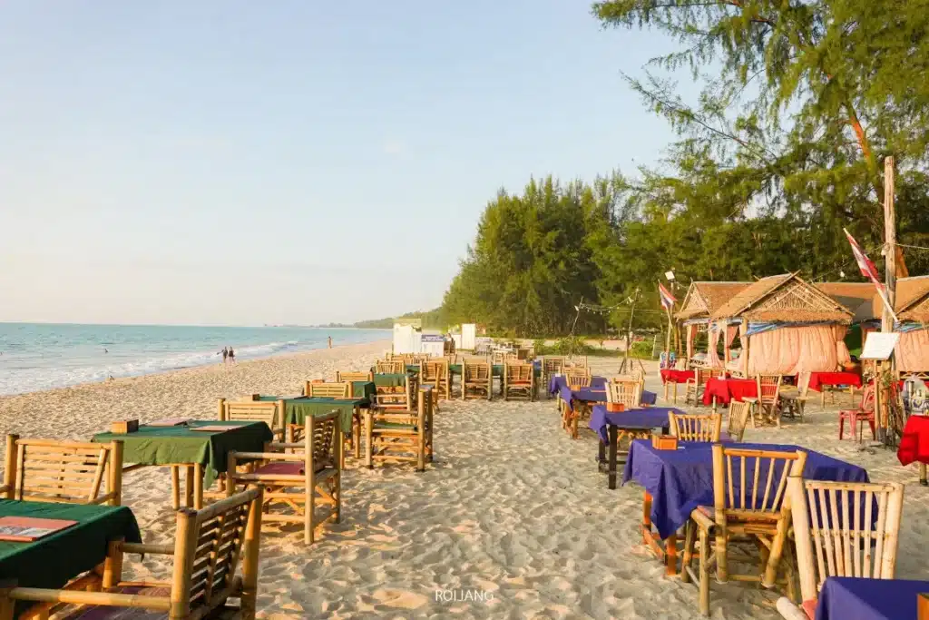 แถวของโต๊ะและเก้าอี้ไม้ถูกจัดเรียงไว้บนชายหาดทรายใกล้ทะเลสีคราม โดยมีกระท่อมเล็กๆ และต้นไม้เป็นฉากหลัง.