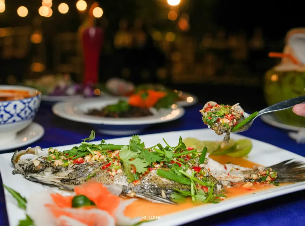 ปลาทั้งตัวปรุงสุกจากบลูซี ตกแต่งด้วยผักชีสับ พริก และซอส เสิร์ฟบนจานขาว ขณะที่ช้อนยกส่วนหนึ่งขึ้นมาให้เห็น อาหารจานอื่น ๆ สามารถมองเห็นได้ในฉากหลัง.