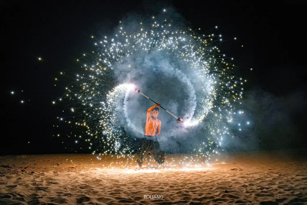 บุคคลหนึ่งทำการเต้นรำกับไฟบนพื้นทรายในยามค่ำคืน สร้างประกายไฟและแสงสว่างเป็นวงกลมขนาดใหญ่ท่ามกลางฉากหลังของทะเลสีคราม.
