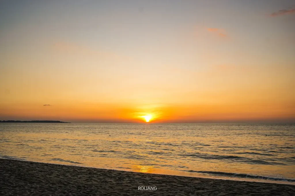 พระอาทิตย์ตกดินเหนือทะเลสีฟ้าสงบ ส่งแสงสีส้มและเหลืองสว่างไสวไปทั่วท้องฟ้าและผืนน้ำ พร้อมชายหาดทรายอยู่เบื้องหน้า.