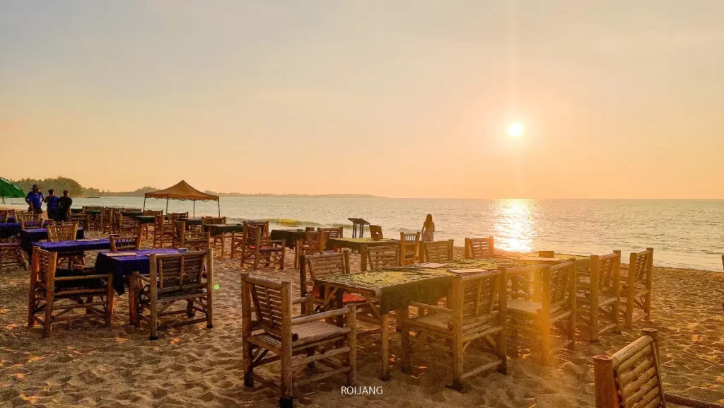 โต๊ะและเก้าอี้ไม้ถูกจัดเรียงบนชายหาดทรายในยามพระอาทิตย์ตกดิน โดยมีดวงอาทิตย์อยู่ต่ำเหนือทะเลสีฟ้าสงบ และมีผู้คนเพียงไม่กี่คนอยู่เบื้องหลัง.