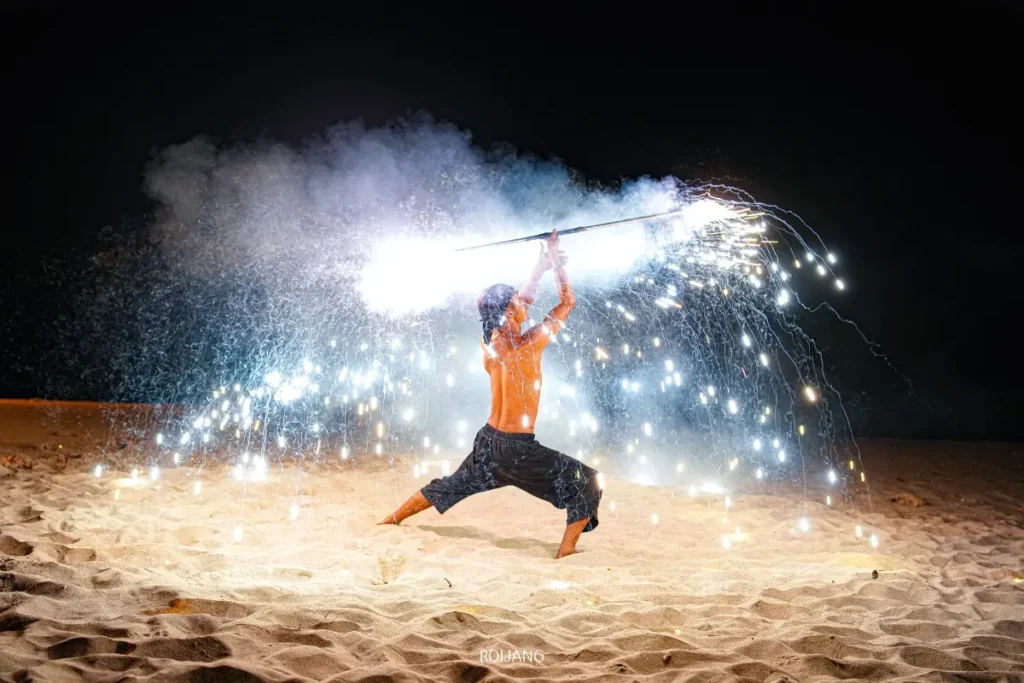 บุคคลหนึ่งกำลังแสดงบนทรายในเวลากลางคืนใกล้ทะเลสีคราม ถือไม้เท้าที่ปล่อยประกายไฟและควันออกมา สร้างการแสดงแสงที่น่าตื่นตาตื่นใจบนฉากหลังที่มืดมิด.