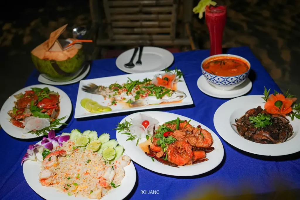 โต๊ะที่จัดเตรียมอาหารทะเลไทยหลากหลายชนิด รวมถึงปลาเผา ปู กุ้ง ข้าวผัด ซุป และเครื่องดื่มสีแดงบนผ้าปูโต๊ะสีน้ำเงิน ชวนให้นึกถึงรสชาติสดใหม่ของทะเลสีคราม.